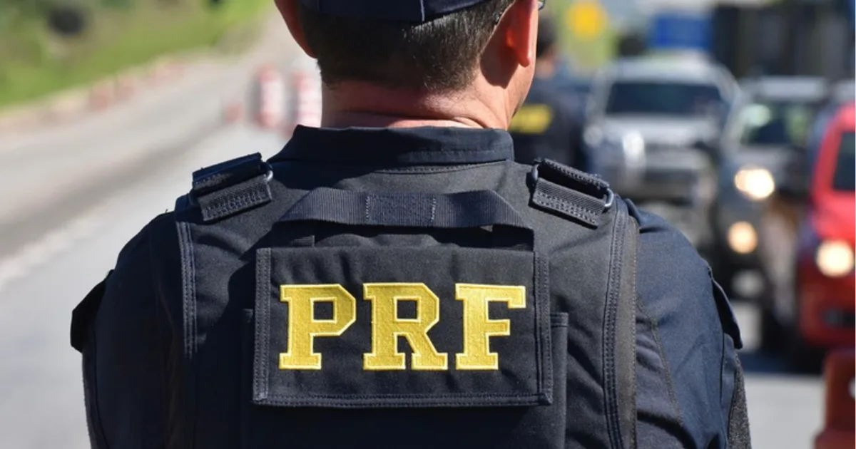 Agente da Polícia Rodoviária Federal de costas com colete preto escrito PRF em amarelo, em rodovia com carros ao fundo.